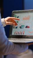 Back view of business woman analysing financial statistics holding laptop standing in start-up office late at night overworking. Busy focused employee using modern technology network wireless overtime photo