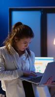 Focused manager woman checking business reports standing in start up office late at night holding laptop. Busy employee using technology network wireless doing overtime reading writing, searching photo