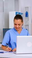 Nurse taking notes on clipboard after reading medical raports of patient from laptop. Healthcare physician in uniform, doctor assistant working in hospital checking registration, writing treatments photo