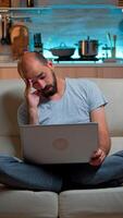Tired man in pajams browsing on internet lifestyle information using laptop computer sitting on sofa. Freelancer working at online project in front on television late at night in kitchen photo