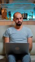 Focused man sitting on sofa and writing online project on laptop late at night in living room. Caucasian male working from home while watching tv, browsing on internet using network technology photo