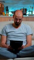 Tired man sitting alone on couch while browsing on internet using tablet computer with modern technology wireless. Caucasian male in pajamas relaxing in front of televion late at night in kitchen photo