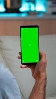 Relaxed man looking at smartphone with mock up green screen chroma key display while relaxing on sofa. Caucasian male holding in horizontal mode phone with isolated display late at night in kitchen photo
