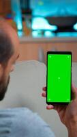 Caucasian male reading online social information on phone with mock up green screen chroma key display. Tired man sitting on sofa using isolated smartphone with technology internet photo