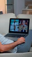 Tired man in pajamas sitting on sofa falling asleep while having online business videocall with teammates using laptop computer. Caucasian male on conference internet web communication photo