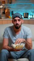 Portrait of man with beard holding popcorn bowl while watching entertainment movie on television. Concentrated person with eye sleep mask sitting on sofa late at night in kitchen photo