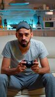 Focused man holding joystick while sitting in front of television winning online games competition. Caucasian male with sleep mask lying on couch making winner gesture late at night in kitchen photo