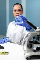 Biochemist researcher woman holding petri dish analyzing blue solution working at scientific experiment in biochemistry hospital laboratory. Biologist scientist doctor examining microorganism sample photo