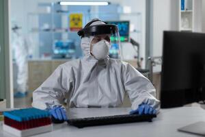 Scientist woman with protection medical equipment against covid19 typing covid19 test results working at vaccine development in scientific hospital laboratory. Virus prevention experiment photo