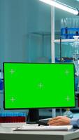 Scientist sitting at desk working on personal computer with mock-up green screen. In background man lab researcher discussing with doctor about vaccine developent bringing blood samples photo