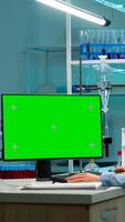 Biochemist sitting at workplace in laboratory using green mock-up screen personal computer with chroma key monitor. coworker working in background of pharmaceutical research centre. photo