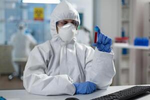 Chemist researcher with protective suit against covid19 holding medical vacutainer analyzing blood results during coronavirus vaccine development. Scientist woman working in hospital lab photo