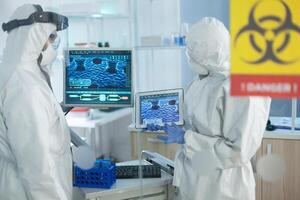 Medical teamwork of scientists wearing protective suit analyzing virus development on tablet during clinical experiment in microbiological hospital laboratory. Danger sign on glass photo