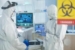 Medical teamwork of scientists wearing protective suit analyzing virus development on tablet during clinical experiment in microbiological hospital laboratory. Danger sign on glass photo