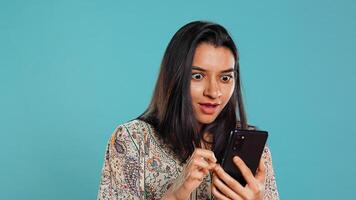 Woman happily scrolling on phone touchscreen to check social media feed, amazed by photo. Indian person browsing internet websites, astonished by content, studio background, camera B photo