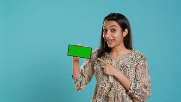 Smiling woman showing isolated screen mobile phone, doing promotion, studio background. Upbeat indian person holding copy space mockup cellphone used for advertising brands, camera A photo