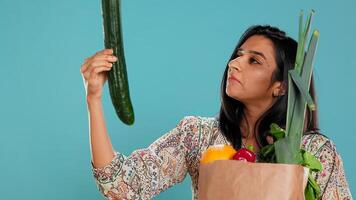 Woman with paper bag with vegetables testing quality, looking at cucumber, isolated over studio background. Person inspecting groceries after buying them from zero waste shop, camera B photo