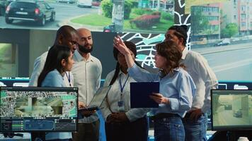 Diverse employees group discussing ways to speed up order delivery, looking for routes to avoid traffic on satellite navigation. Agency operators use CCTV footage in monitoring room. Camera A. photo
