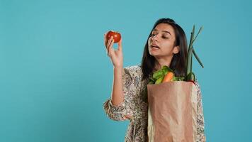 Woman with recycled paper bag in hands filled with vegetables testing quality, studio background. Sustainable living person inspecting groceries after doing purchases in shop, camera A photo
