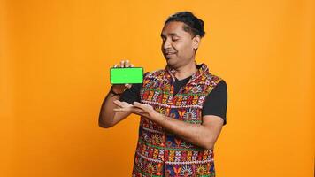 Content creator presenting green screen mobile phone, isolated over studio background. Indian person reviewing copy space mockup smartphone device for internet channel, camera B photo