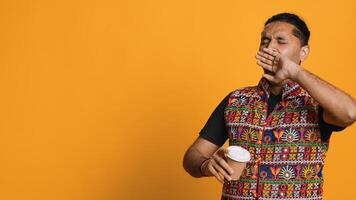 Sleepy man yawning and rubbing eyes. sipping coffee to wake up and gain energy. Exhausted person feeling fatigued after sleepless night, drinking caffeinated beverage, studio backdrop, camera A photo