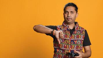 Upset man showing thumbs down sign gesturing holding controller, disapproving of videogames. Displeased indian person against gaming, doing rejection hand gesture, studio background, camera A photo