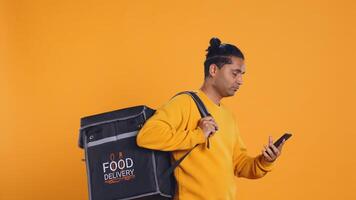 Man holding thermal backpack answering phone, fulfilling clients orders, studio backdrop. Food delivery service worker doing delivery to customers, talking on smartphone, camera B photo