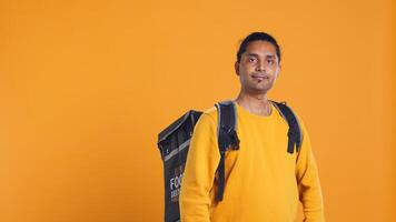 Portrait of confident indian man holding thermal backpack, crossing arms, studio background. Radiant nonchalant delivery person prepared to deliver takeaway food to customers, camera B photo