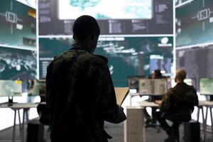 Infantry unit soldier overseeing intelligence gathering activity in military command center, comparing the data between case files and big screen. Army conscript examines satellite information. photo