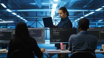 Middle eastern manager in server hub workplace doing inspection on electronics and multiracial workers. Team leader reading paperwork on laptop, verifying infrastructure documents, doing audit photo