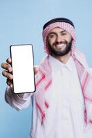 Smiling muslim man presenting smartphone empty screen with copy space for software app promotion. Cheerful person in arabic clothes showing mobile phone with blank display closeup photo