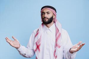 Confused muslim man in traditional attire spreading hands and expressing uncertainty portrait. Puzzled arab person shrugging shoulders with open arms and looking at camera photo
