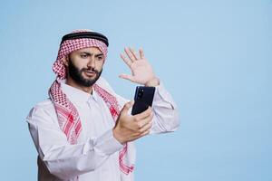 Muslim man in traditional thobe and headscarf speaking in chat on smartphone and waving hi in studio. Arab person greeting and chatting in videoconference on mobile phone photo
