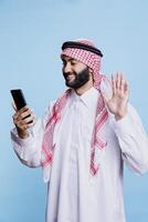 Muslim man greeting with friendly hand wave and smile while chatting in videocall on smartphone. Cheerful arab in thobe saying hello while speaking in online meeting using mobile phone photo