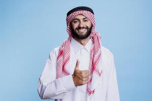 Smiling muslim man in white thobe and checkered headdress showing thumb up studio portrait. Cheerful arab posing with positivity and satisfaction gesture and looking at camera photo