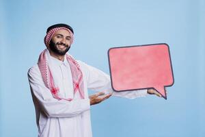 Cheerful man dressed in muslim traditional clothes showing at blank red dialog banner and looking at camera. Smiling carefree arab holding speech bubble mockup with copy space photo