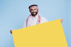 Smiling muslim man dressed in traditional clothes holding blank yellow banner with copy space studio portrait. Arab person wearing cultural attire standing with empty colorful board photo