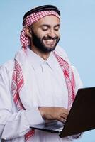 Smiling man wearing islamic thobe and ghutra texting in social media while standing with laptop in studio. holding portable computer and chatting online with cheerful expression photo