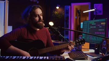 Songwriter producing tunes on acoustic guitar in his personal studio, using a microphone to sing the chords. Audio engineer works with soundboard and equalizer to compose new music. Camera B. photo