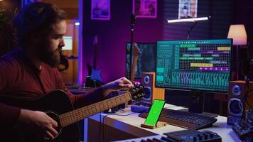 Artist learning to tune his acoustic guitar with app tutorial, using greenscreen display on smartphone an practicing music lessons. Young musician doing rehearsal with instrument. Camera B. photo
