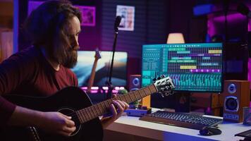 Artist playing guitar and uses DAW software interface to create music, looking at mastering controls on computer screen. Musician recording soundtracks with tools, acoustical engineering. Camera B. photo