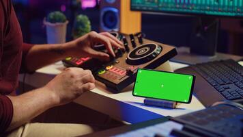 Musical performer creating mixed sounds and tunes with mixing console, greenscreen running on phone display. Artist recording new notes using equalizer and daw software on computer. Camera A. photo
