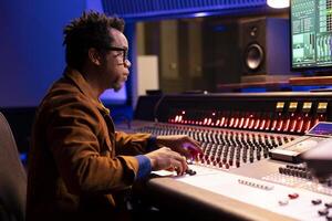 African american sound designer uses control desk with automatic equalizer, mixer and faders to edit music in post production. Audio expert processing audio recordings, mixing console. photo