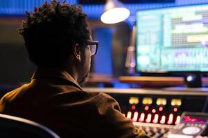 African american record producer mixing and mastering tracks with digital software, working with motorized faders and knobs to adjust sound settings. Audio engineer producing new music. photo