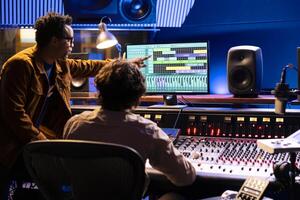 Team of artists collaborating on processing recorded files with audio software, pushing faders to adjust volume levels. Technician and singer creating music on control panel board in studio. photo