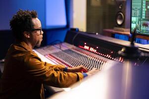 Audio expert processing music from soundproof vocal booth at professional studio, recording songs and editing them after in post production. Technician uses mixing console and buttons or sliders. photo