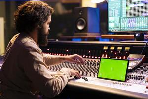 Professional music engineer recording his sounds next to tablet with mockup, editing in control room using faders and knobs to do mix and master. Young producer creating new tracks for an album, photo