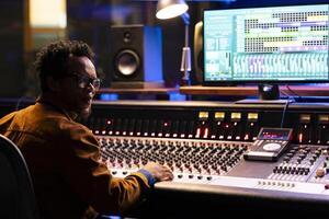 Sound designer working on control desk in professional studio, using faders and switchers to edit and produce new music for his album. Young producer creating tracks, mix and master techniques. photo
