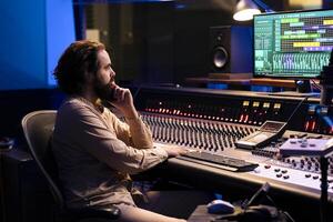Tracking engineer uses audio console in professional recording studio, adjusting volume levels while pressing buttons on panel board. Skilled audio technician operates mixing and mastering gear. photo
