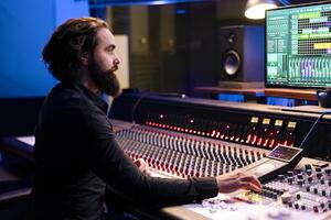 Tracking engineer editing new music with digital audio software and mixing console in control room, working in post production at studio. Audio technician operates sliders and knobs. photo